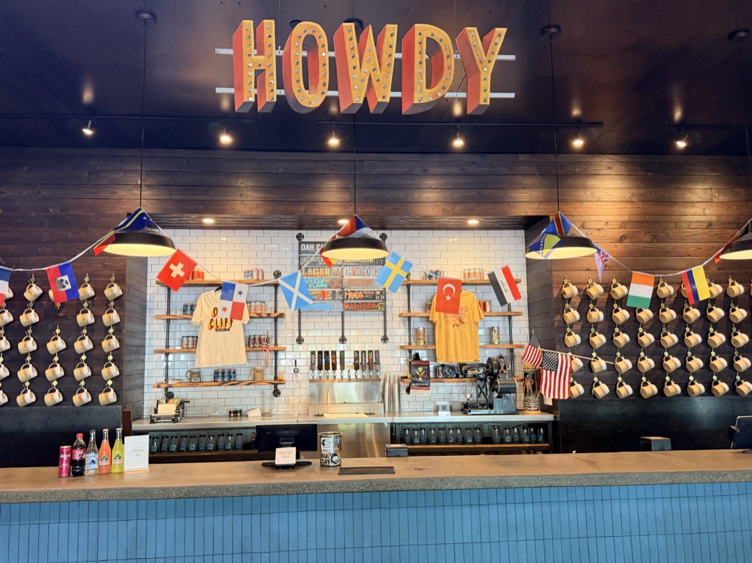 Oak Cliff Brewing’s taproom bar beneath a large HOWDY sign, with colorful flags and hanging mugs.