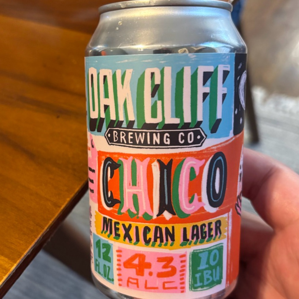 A hand holding a colorful Chico Mexican Lager can beside a wooden table.